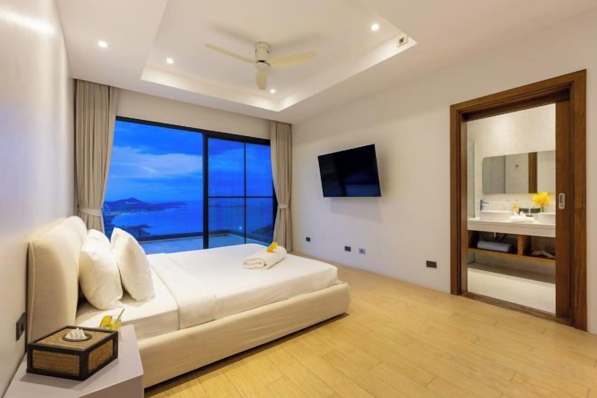 A modern hotel room with a large bed, a flat-screen TV, a balcony with a view, and a wooden door.