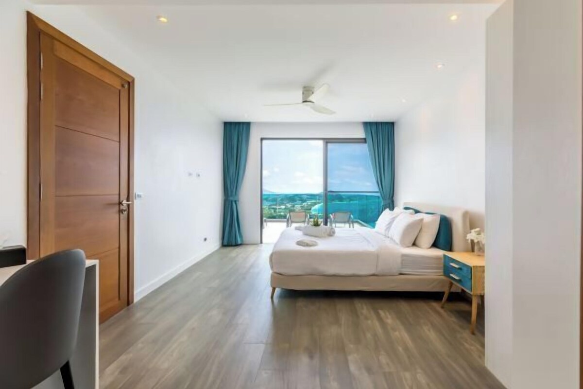 Une chambre à coucher comprenant un lit, un bureau, une chaise, un ventilateur de plafond et une grande fenêtre avec des rideaux.
