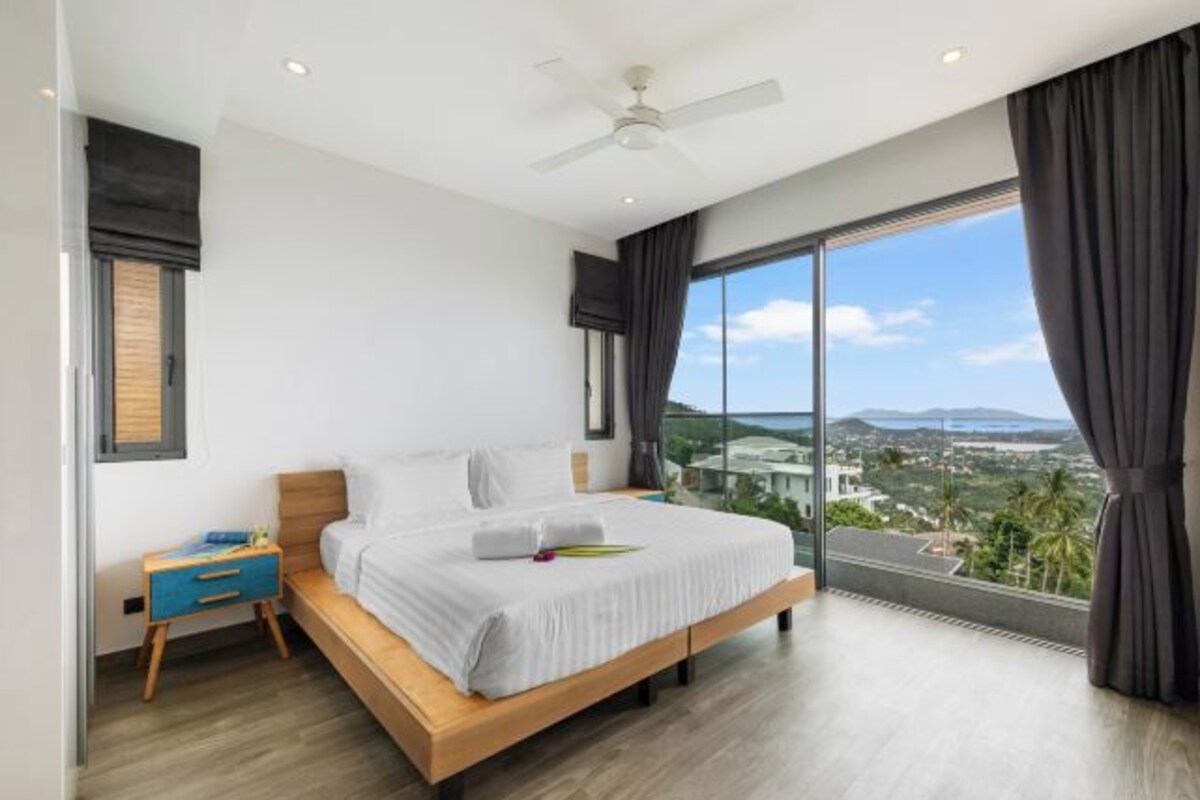 Une chambre à coucher comprenant un lit, une table de chevet, un ventilateur de plafond et une grande fenêtre donnant sur un beau paysage.