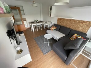 A modern living room with a grey sofa, a small white table, a dining area with a white table and chairs, and a wooden accent wall.