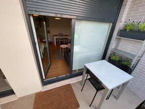 En liten uteplats med ett bord och stolar, en skjutdörr i glas som leder in till en matplats inomhus, och en tegelvägg med en hylla för växter.