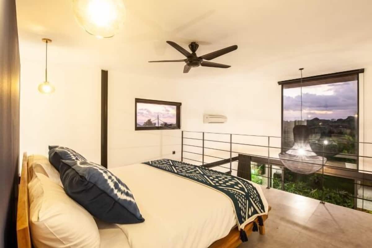 Un dormitorio moderno con cama, ventilador de techo, un cuadro en la pared y balcón con vistas.