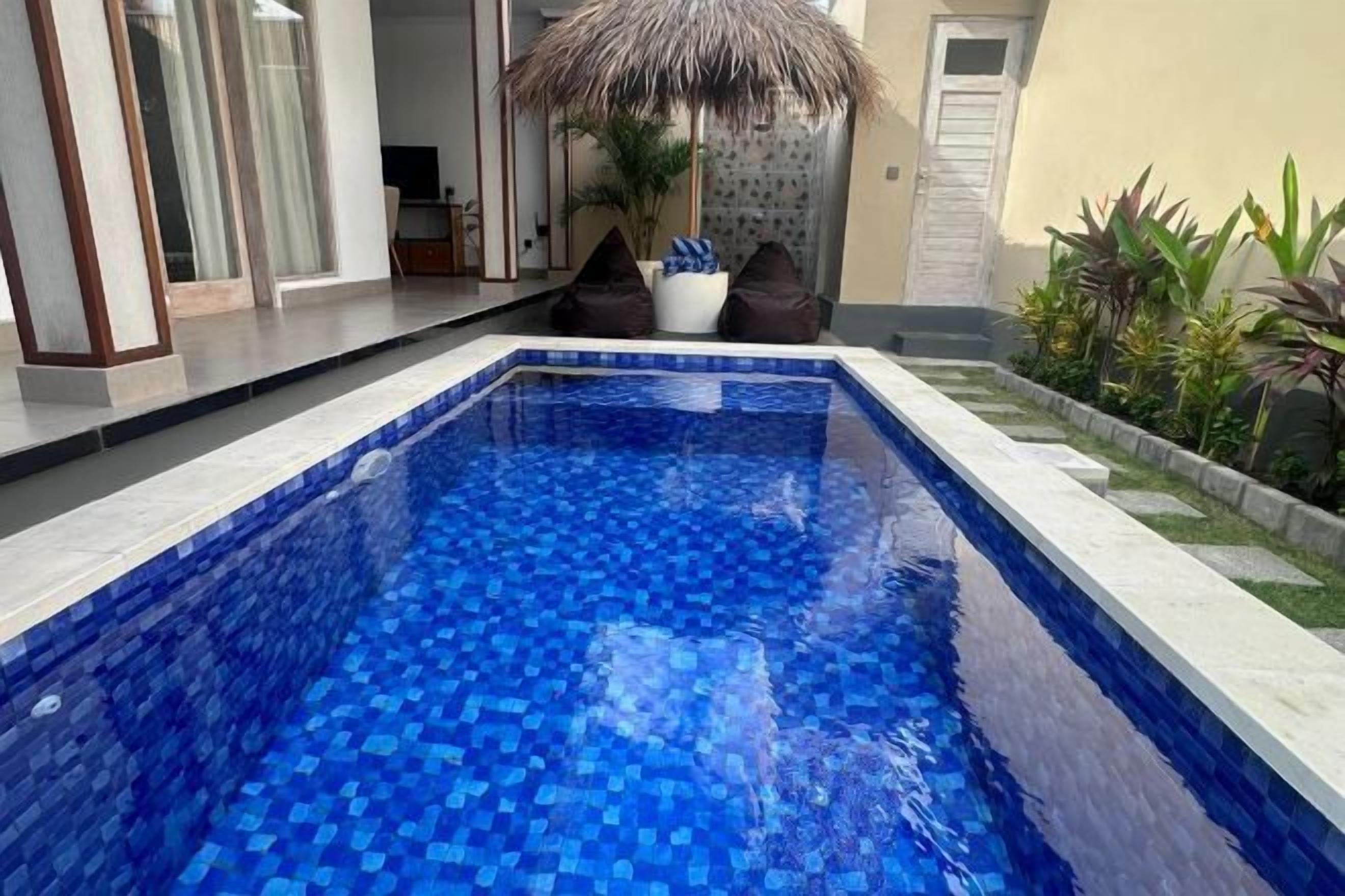 A swimming pool with blue mosaic tiles, a thatched umbrella, and a potted plant.