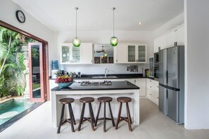 A modern kitchen with a central island, bar stools, a refrigerator, and a view of a pool. - Villa Annecy, Fully Staffed Luxury Villa Central Seminyak (Bali)