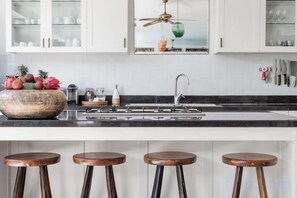 A kitchen with a black countertop, white cabinets, a large bowl of fruit, a ceiling fan, and three stools.