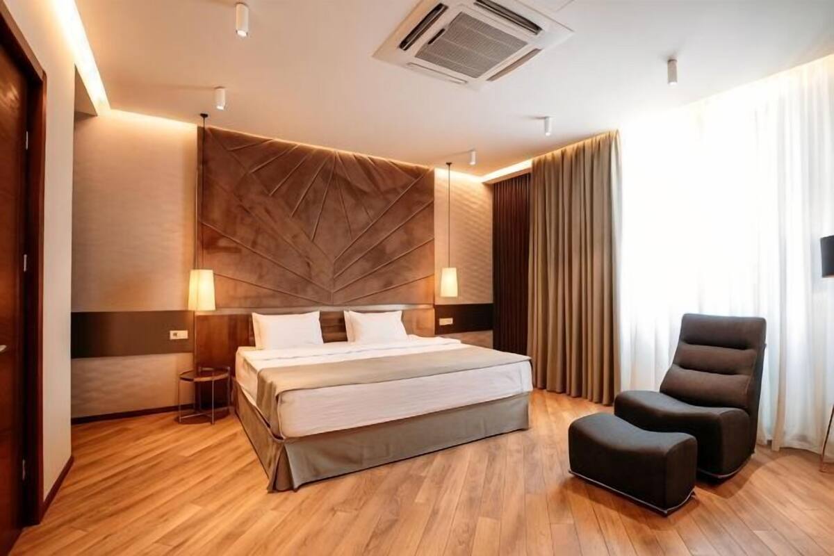 A modern hotel room with a large bed, a bedside table, a chair, and a headboard with a geometric pattern.