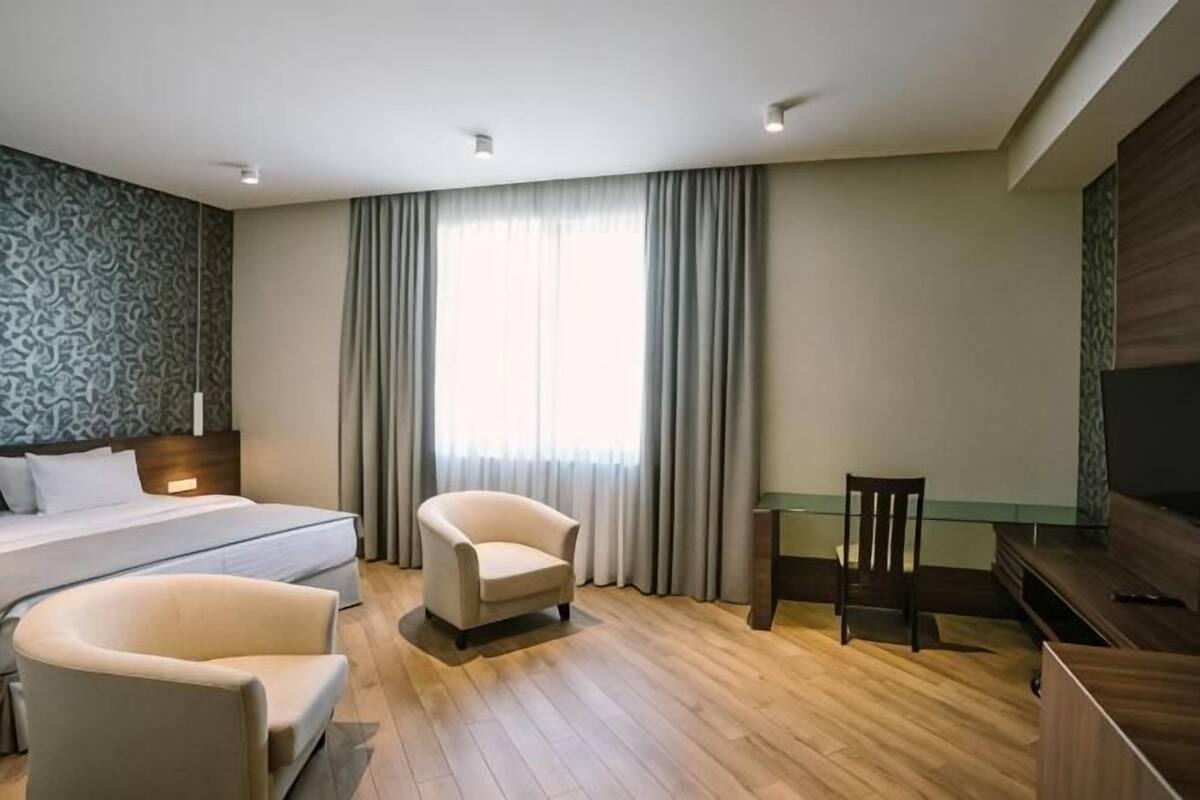 A hotel room with a bed, two armchairs, a TV, and a large window with curtains.