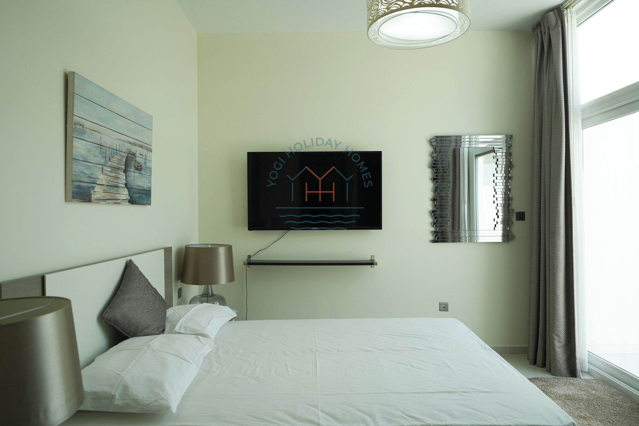 A hotel room with a bed, a TV mounted on the wall, a painting on the wall, and a window with curtains.