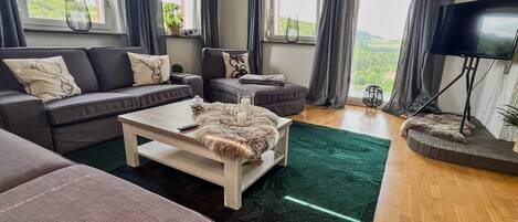Un salon moderne avec un canapé, un pouf et une table basse. On y trouve également un téléviseur et une plante.