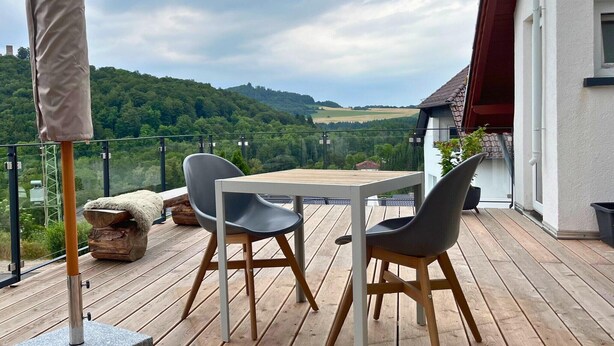 Une terrasse en bois avec une table et des chaises, offrant une vue panoramique sur un paysage pittoresque composé de collines et d’un ciel dégagé.