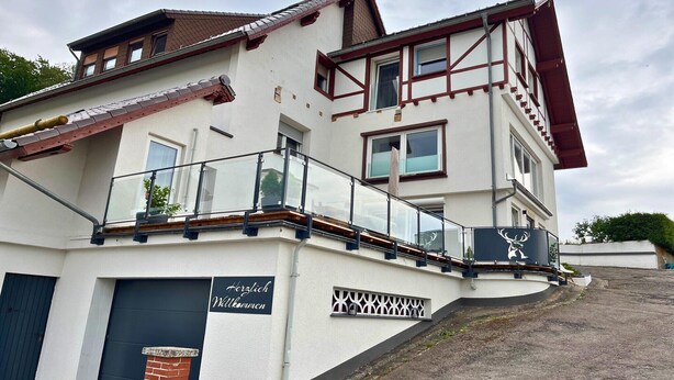 A two-story building with a balcony, a sign reading "Herzlich Willkommen," and a deer logo. - Guest rooms Eifelglück in Pelm (Pelm)