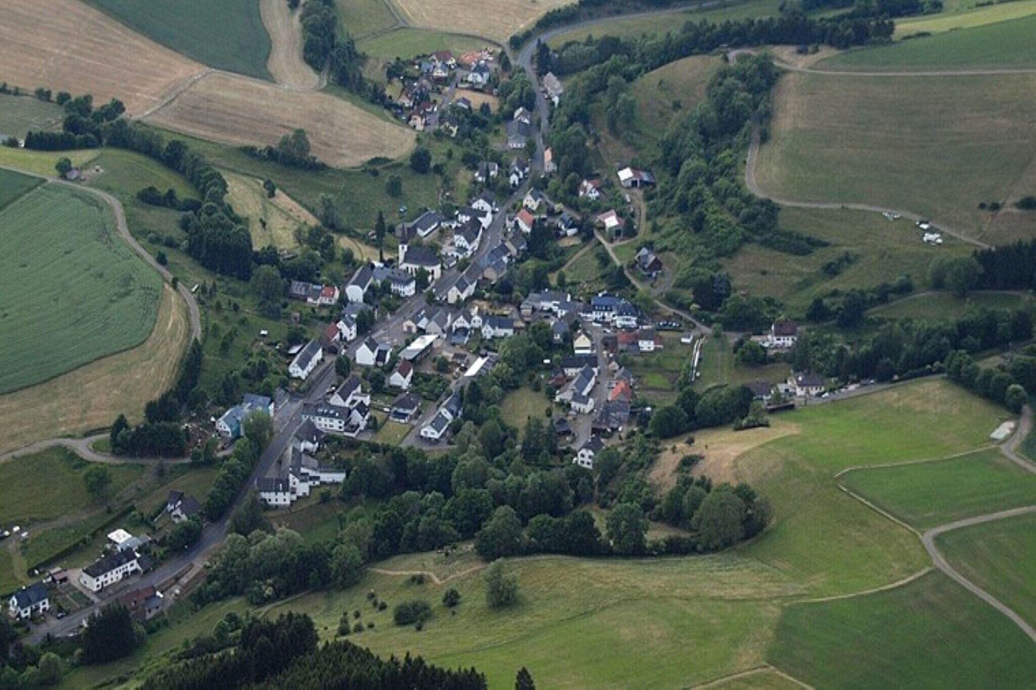 Vista aérea de un pequeño pueblo con casas agrupadas alrededor de una calle central, rodeado de campos.