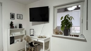 A room with a white desk, a chair, a TV mounted on the wall, a window with blinds, and a potted plant on the windowsill. - Guest rooms Eifelglück in Pelm (Pelm)