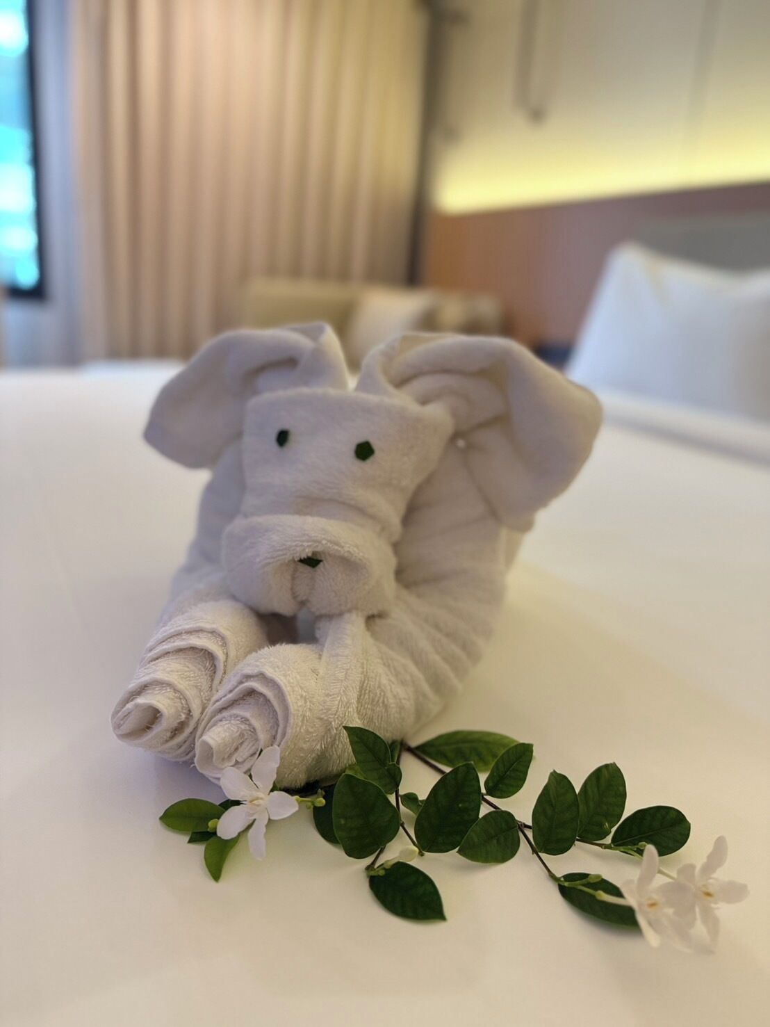Ein ordentlich gefaltetes Handtuch in Form eines Hasen auf einem Bett mit weißer Bettwäsche, begleitet von einem kleinen Zweig mit grünen Blättern und weißen Blüten.