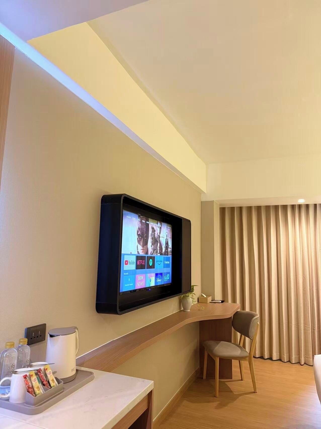 Une chambre d’hôtel équipée d’un téléviseur à écran plat fixé au mur, qui permet d’accéder à divers services de diffusion en continu, d’une petite table avec une machine à café et quelques en-cas, d’une chaise et d’une fenêtre avec des rideaux.