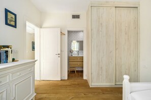 A bedroom with a white bed, a wooden floor, a white wardrobe, and a bathroom with a tiled wall and a small stool. - MAMAD - Rosh HaNikra Garden Villa by Sea N' Rent (Rosh HaNikra)