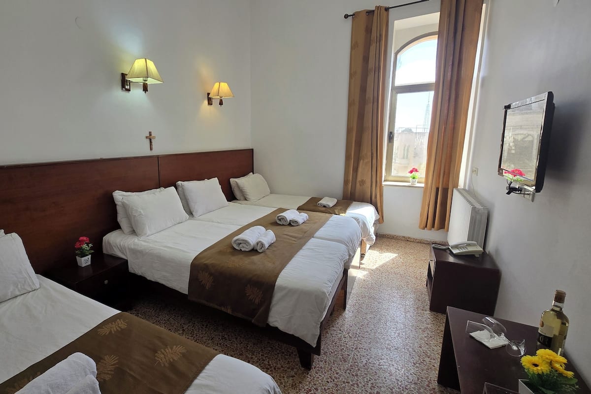 A hotel room with two beds, a wooden headboard, a small table with a telephone, a window with curtains, and a wall-mounted TV.