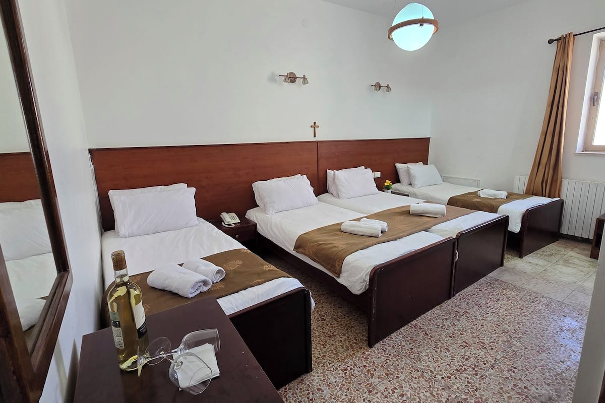 A hotel room with two beds, a wooden headboard, a small table with a bottle and glasses, and a window with curtains.