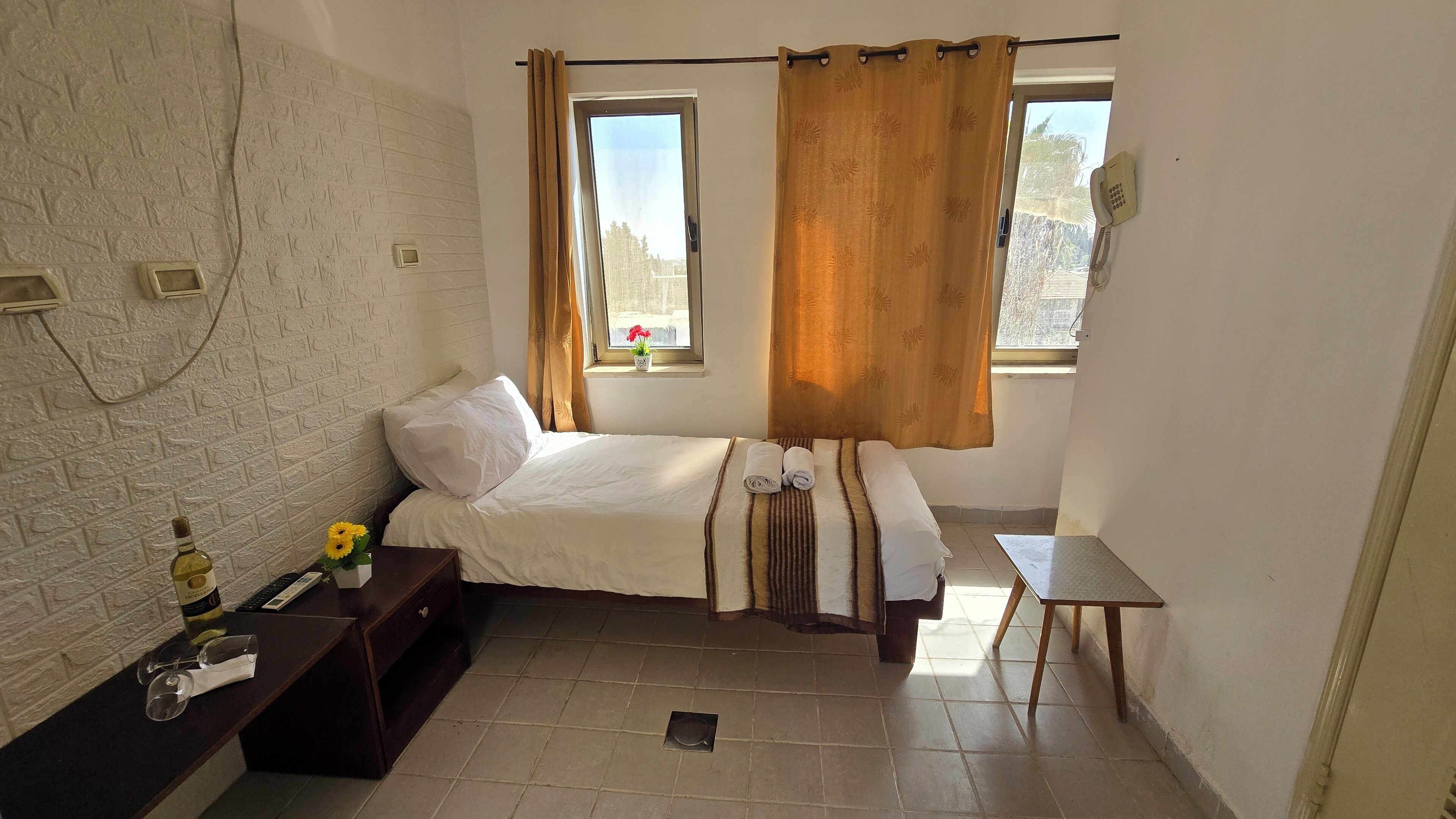 A small, tidy room with a bed, a small table, a telephone, and a window with curtains.