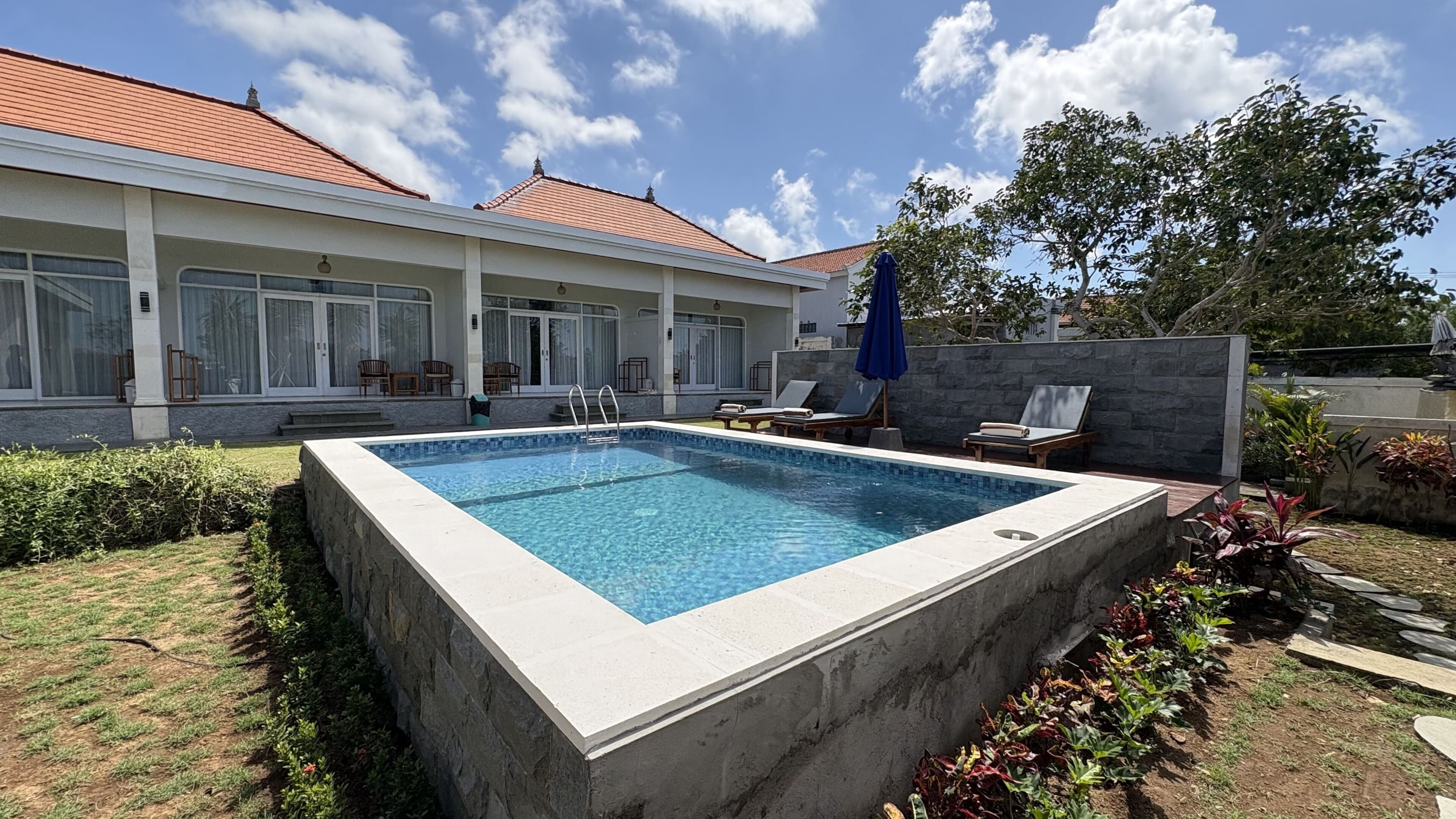 Ein rechteckiger Pool mit einer gefliesten Terrasse, umgeben von einem Garten mit verschiedenen Pflanzen und einer Steinmauer.