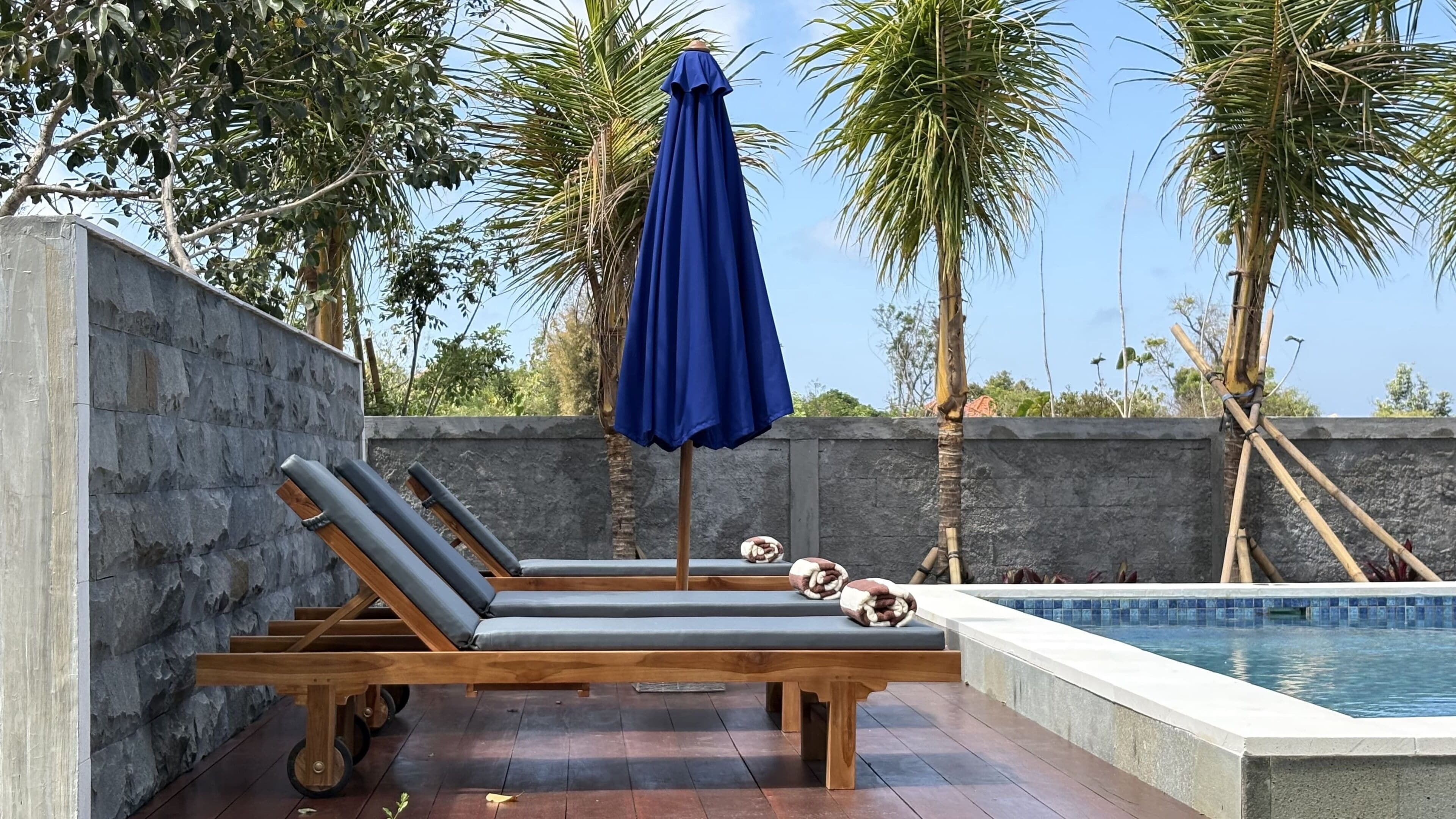 A poolside area with lounge chairs, a blue umbrella, and a stone wall.