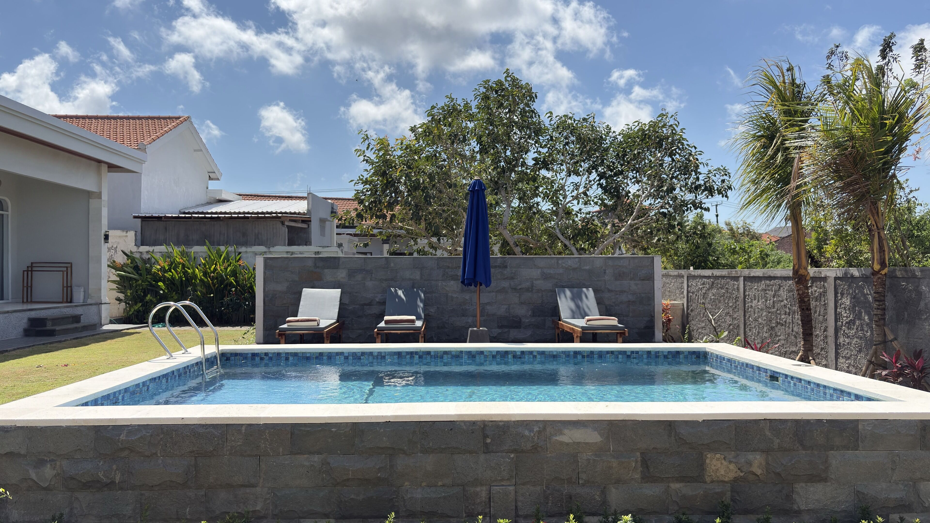 A rectangular swimming pool with a stone wall, two lounge chairs, and a blue umbrella.