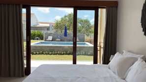 A hotel room with a large bed, a view of a swimming pool, and a patio area with chairs and a blue umbrella. - Octa House (Kutuh)