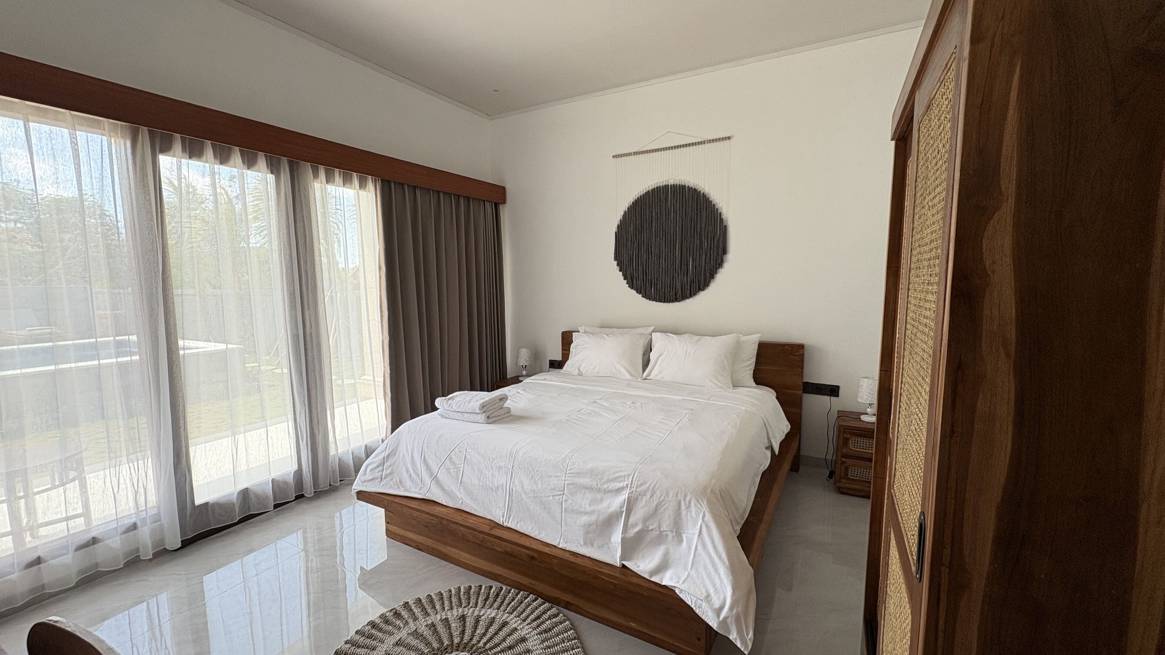 A bedroom with a large bed, a round wall decoration, and a view through sheer curtains.