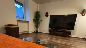 Ein modernes Wohnzimmer mit einem großen Flachbildfernseher, einer Topfpflanze und einem Fenster mit Blick auf Gebäude.