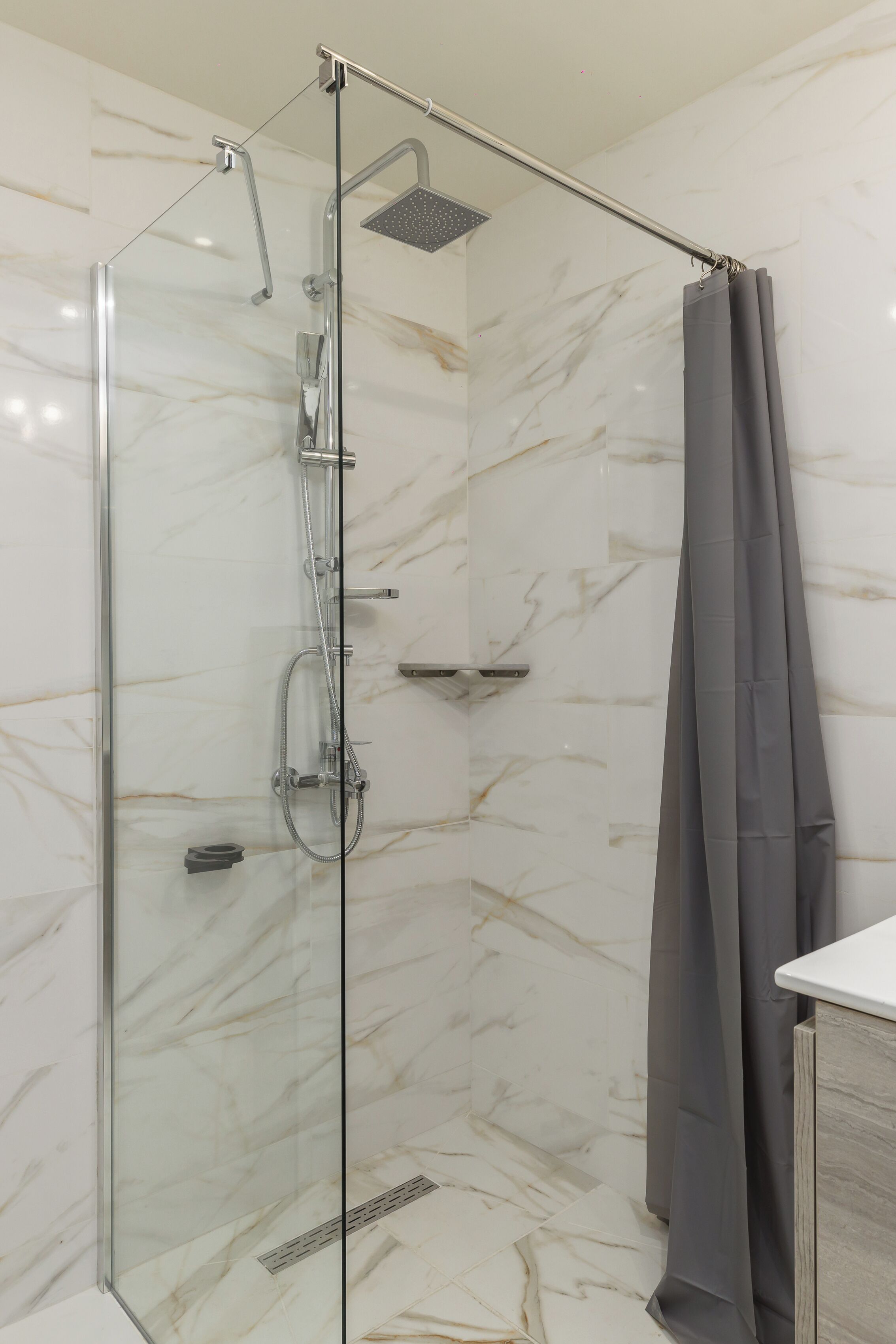 A glass-enclosed shower with a handheld showerhead and a rain shower head, a grey shower curtain, and a marble-patterned wall.