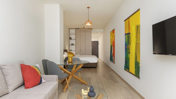 Un salon moderne avec une table basse en verre, un canapé agrémenté de coussins colorés, un coin repas avec une table et des chaises en verre, et des œuvres d’art accrochées aux murs.