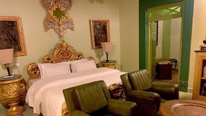 A hotel room with a large bed, two green leather chairs, a round wooden table, and ornate gold accents. - Partap Mahal (Bikaner)