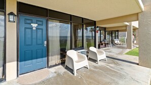 A hotel entrance with a blue door labeled "Shell Room 102," a covered patio with two white chairs, and a view of the outdoors.