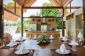 Un comedor con una mesa larga dispuesta para una comida, una cocina al fondo y vistas a árboles.