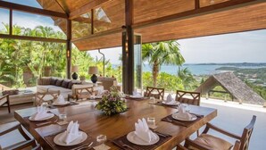 A spacious dining area with a wooden table set for a meal, overlooking a lush landscape with palm trees and a view of the sea.