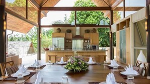 A dining area with a central table set for a meal, surrounded by chairs, and a view of a kitchen and outdoor greenery.