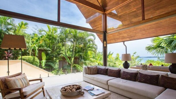 A modern living room with a large window, a sofa, a coffee table, and a view of lush greenery and the sea.