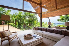 A modern living room with a large window, a sofa, a coffee table, and a view of lush greenery and the sea. - Panacea Retreat 4 Bedroom - Kalya Residence (Koh Samui)