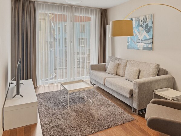 A modern living room with a sofa, a coffee table, a lamp, and a view through sheer curtains.