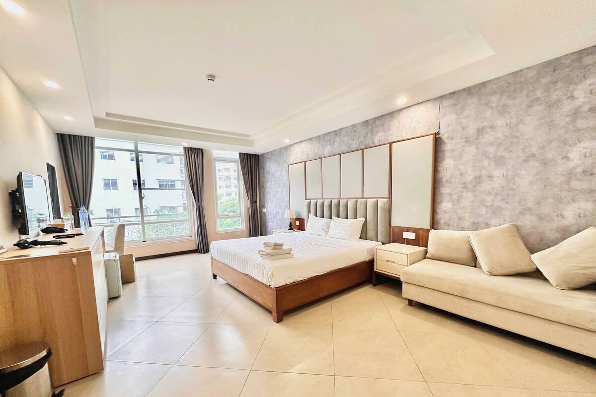 A modern hotel room with a large bed, a sofa, a desk, and a view of the outside.