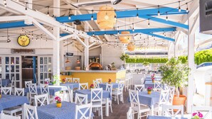 An outdoor restaurant with white tables and chairs, blue and white checkered tablecloths, hanging lights, and a pizza oven. - Collins Holiday (Sianożęty)