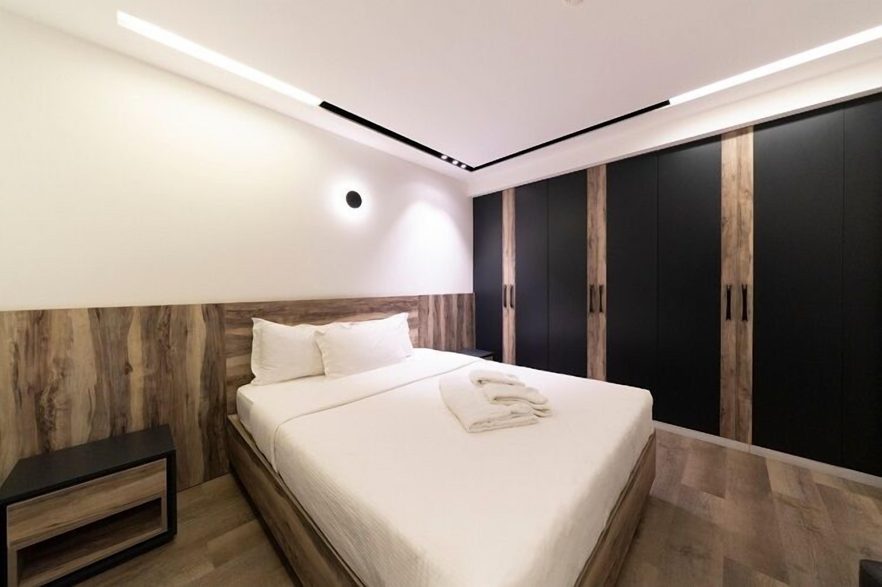 A modern bedroom with a wooden headboard, a large bed with white linens, a nightstand, and a wardrobe with dark doors.