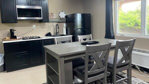 Een kleine keuken met zwarte kastjes, een magnetron en een spoelbak. Er staat een eettafel met stoelen en een raam met uitzicht op groen.
