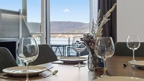 A dining table set with plates, cutlery, and wine glasses, with a view of a body of water and mountains in the background. - Maria Blick Lake View Apartments (Maria Woerth)
