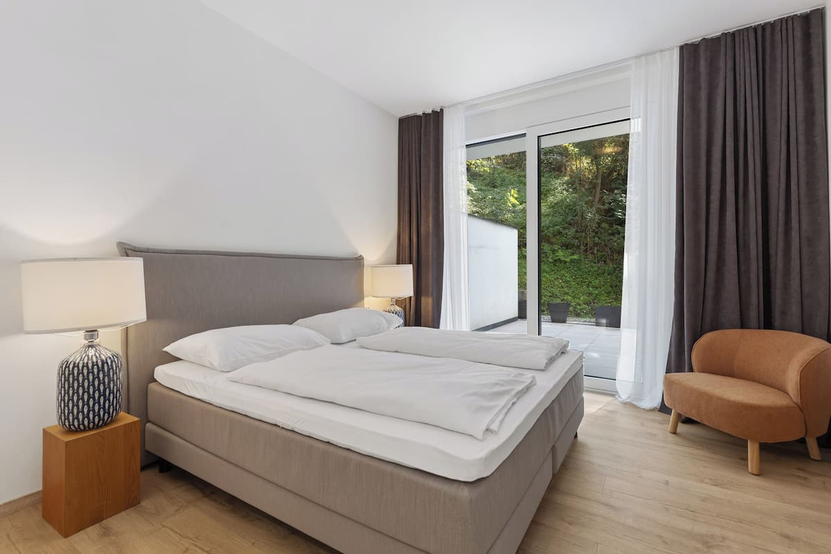 A modern hotel room with a large bed, a bedside table with a lamp, a chair, and a view of greenery through the window.