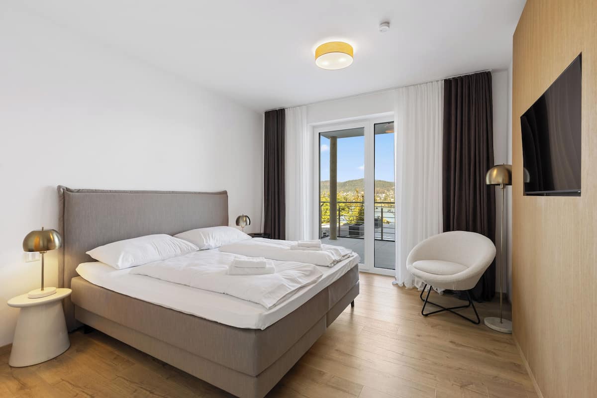 A modern hotel room with a large bed, a chair, a nightstand, and a view of the outdoors.