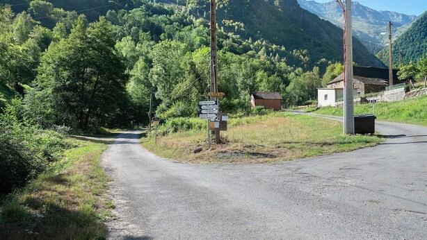 Une route de campagne, bordée d’un chemin de gravier menant à un petit bâtiment, entouré d’une végétation luxuriante et de montagnes.