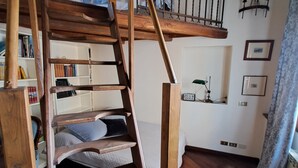 Une pièce de style « loft », avec une échelle en bois menant à un lit superposé. On y trouve une bibliothèque, un bureau avec un ordinateur et un radiateur fixé au mur.