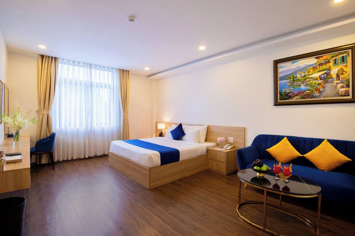 A hotel room with a wooden bed, a blue sofa, a small table with fruit and drinks, and a painting on the wall.