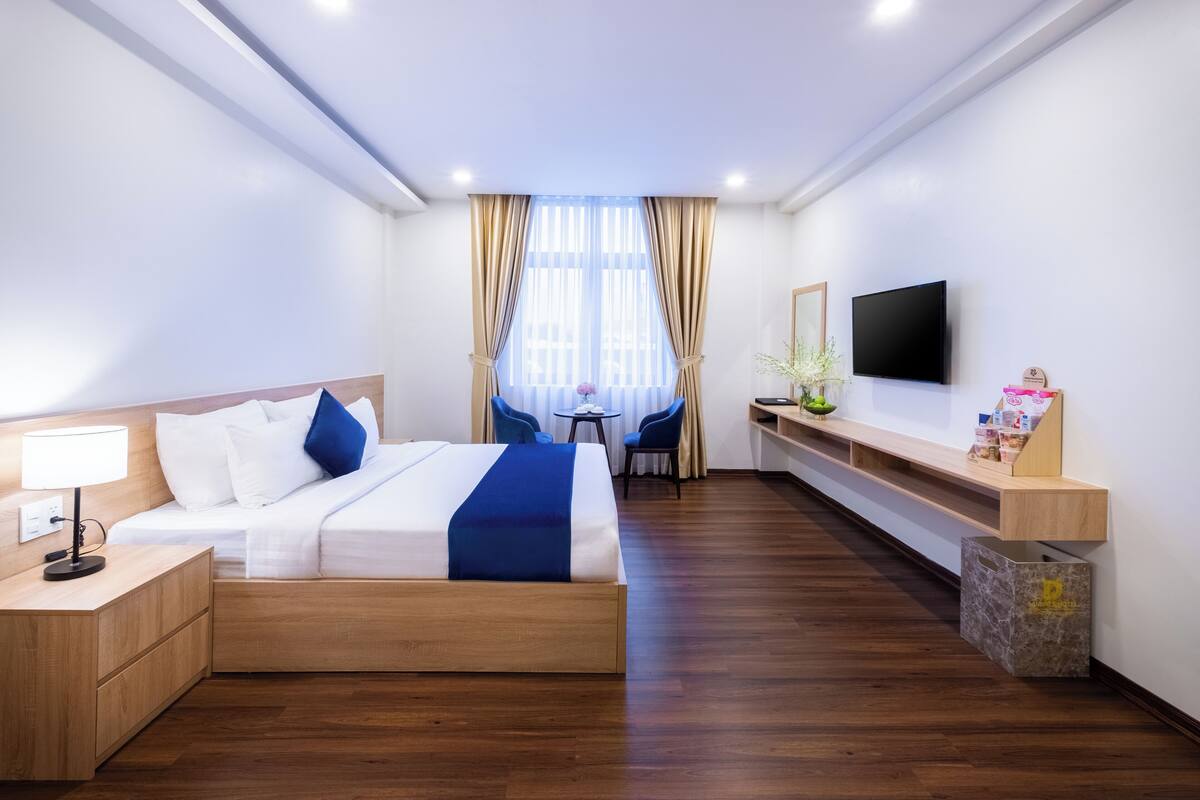 A modern hotel room with a wooden bed, a desk, a chair, a TV, and a small table with a vase.